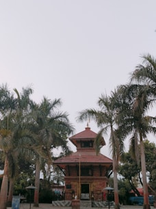A traditional temple structure with a red brick facade and tiered roof is surrounded by tall palm trees. The sky above is clear, creating a serene and calm atmosphere.