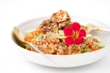 A close-up of a beautifully plated dish featuring a mixture of grains topped with sesame seeds, vibrant red edible flowers, and chopped vegetables. The meal is garnished with pieces of crispy, fried chicken and served in a white bowl with a golden spoon.