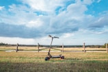 An electric scooter is positioned on a grassy field against a backdrop of a wooden fence and an expansive, partly cloudy sky. The landscape is vast and open, with a mixture of green grass and clear blue skies.