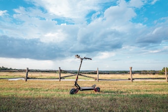 An electric scooter is positioned on a grassy field against a backdrop of a wooden fence and an expansive, partly cloudy sky. The landscape is vast and open, with a mixture of green grass and clear blue skies.