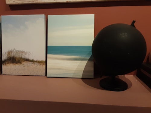 Two framed beach-themed artworks sit on a shelf. The first shows dune grass growing on sandy terrain under a soft sky. The second depicts a serene seascape with a calm ocean meeting the horizon. A dark, spherical object resembling a globe stands beside the images, providing a contrasting element.