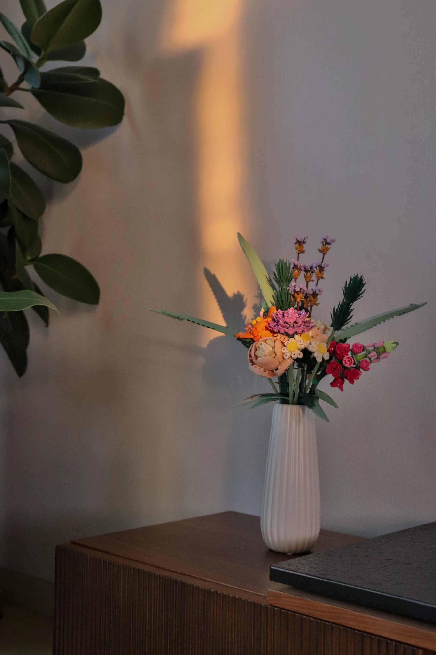 A brick-built flower bouquet in a white ribbed vase on a wooden sideboard in evening light