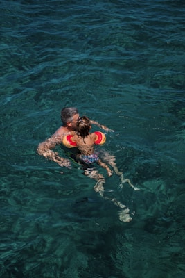 A person is holding a child in the water, both wearing swimsuits. The child has arm floaties for safety. The water is clear, revealing shades of blue and green.