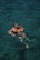A person is holding a child in the water, both wearing swimsuits. The child has arm floaties for safety. The water is clear, revealing shades of blue and green.