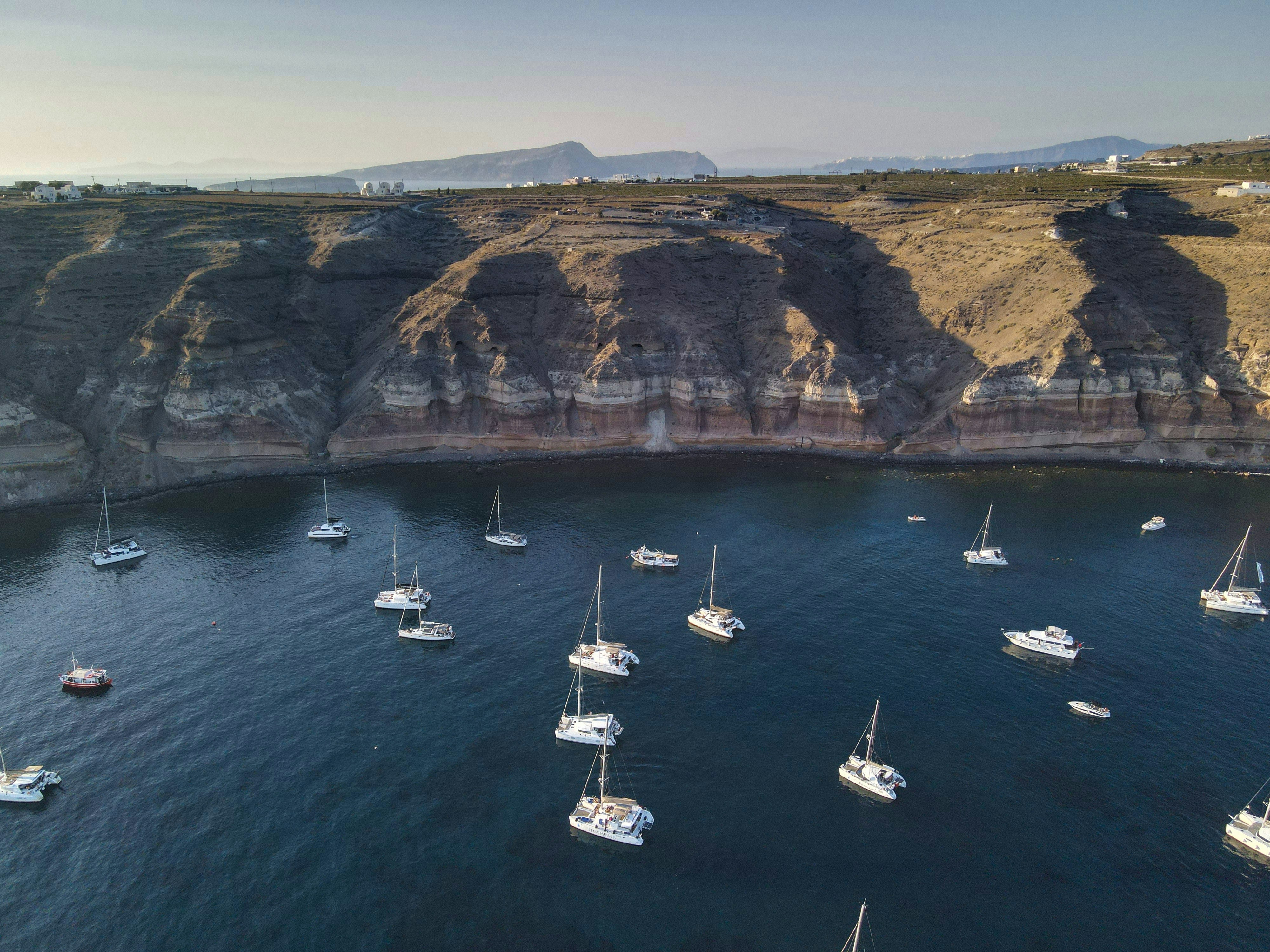 white boats on sea near brown mountain during daytime, A view above one of the most famous beaches in Santorini island, Greece.