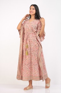 A woman is wearing a long flowing dress with intricate patterns and tassels, standing in a relaxed pose. Her expression is cheerful and she is looking slightly to the side. The dress features a mix of pink and orange hues with decorative borders.