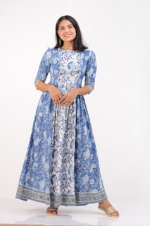 A woman stands wearing a long, flowing blue and white dress with intricate floral patterns. She is smiling and has long dark hair cascading down. Her shoes are open-toe and golden in color, complementing her outfit.