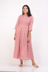 A woman is standing against a plain white background, wearing a long-sleeved, full-length pink floral dress. She is smiling gently and has long dark hair. She is wearing gold slip-on sandals and no other visible accessories.