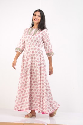 A woman is wearing a long white dress with pink patterns and intricate green detailing around the collar and sleeves. She is posing gracefully against a plain white background, exuding a cheerful expression. Her hair is dark and left open, and she is wearing beige sandals.