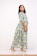 A person is standing and smiling while wearing a long floral dress with a blue and yellow pattern. The dress features large, prominent flower designs with three-quarter-length sleeves. The person is wearing glittery, flat sandals and has medium-length dark hair. The background is plain and white.