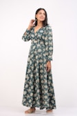 A woman stands confidently wearing a long, patterned dress with shades of teal and cream. The dress has long sleeves and a V-neckline. She is smiling slightly, with one hand touching her hair and the other resting by her side. She wears flat sandals, and her hair is styled straight.