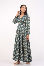 A woman stands confidently wearing a long, patterned dress with shades of teal and cream. The dress has long sleeves and a V-neckline. She is smiling slightly, with one hand touching her hair and the other resting by her side. She wears flat sandals, and her hair is styled straight.