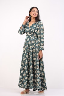 A woman stands confidently wearing a long, patterned dress with shades of teal and cream. The dress has long sleeves and a V-neckline. She is smiling slightly, with one hand touching her hair and the other resting by her side. She wears flat sandals, and her hair is styled straight.
