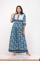 A person stands confidently wearing a long blue floral dress with geometric patterns at the top and tassels near the neckline. The dress has a flowing design, and the person is wearing flat, open-toed sandals, set against a simple white background.