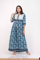 A person stands confidently wearing a long blue floral dress with geometric patterns at the top and tassels near the neckline. The dress has a flowing design, and the person is wearing flat, open-toed sandals, set against a simple white background.