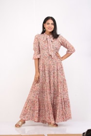 A woman stands confidently with one hand on her hip. She is wearing a long, floral-print dress with a bow at the collar, and her shoes are slip-on sandals. The background is plain and white, enhancing the focus on her attire.
