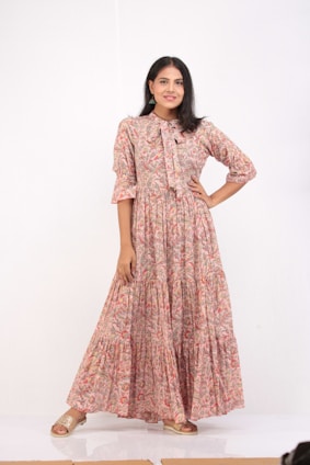 A woman stands confidently with one hand on her hip. She is wearing a long, floral-print dress with a bow at the collar, and her shoes are slip-on sandals. The background is plain and white, enhancing the focus on her attire.