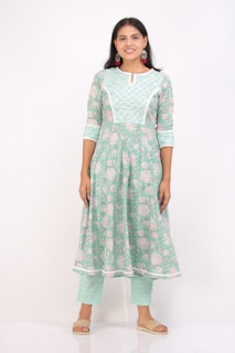 A woman smiles while wearing a floral-patterned, green and white dress paired with matching pants. She is standing against a plain white background and is accessorized with red and silver earrings. Her footwear consists of beige sandals.