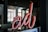 A red neon sign displaying the stylized letters 'al' is prominently featured in the foreground. The sign appears to be situated indoors, with a backdrop of large windows allowing natural light to stream in. The interior setting includes dark walls and a ceiling, along with a reflective surface beneath the neon sign.