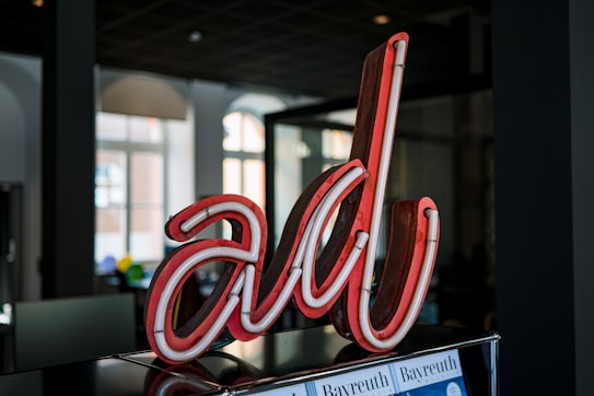 A red neon sign displaying the stylized letters 'al' is prominently featured in the foreground. The sign appears to be situated indoors, with a backdrop of large windows allowing natural light to stream in. The interior setting includes dark walls and a ceiling, along with a reflective surface beneath the neon sign.
