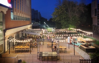 A lively outdoor dining area illuminated by strings of glowing lights. Numerous tables and chairs are arranged neatly on a patio beside a large building. A food truck is parked nearby, adding to the vibrant atmosphere. It's evening, with a deep blue sky providing a backdrop to the trees and cityscape in the distance.