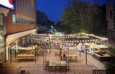 A lively outdoor dining area illuminated by strings of glowing lights. Numerous tables and chairs are arranged neatly on a patio beside a large building. A food truck is parked nearby, adding to the vibrant atmosphere. It's evening, with a deep blue sky providing a backdrop to the trees and cityscape in the distance.