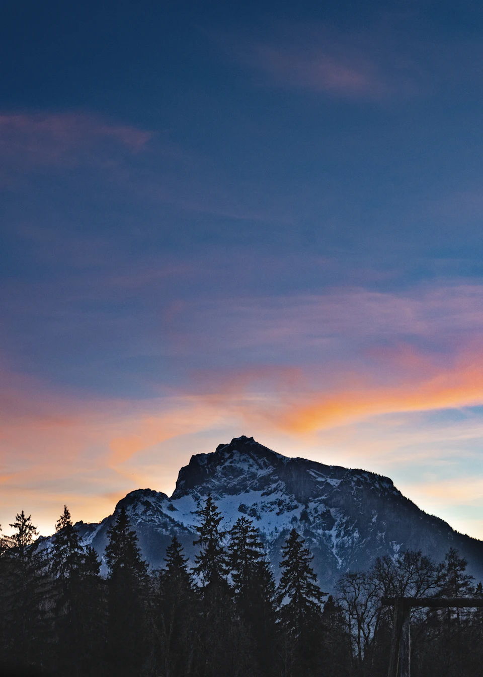 The image depicts a majestic mountain range with snow-capped peaks against a deep blue sky, creating a stunning backdrop for the scene. The mountains are bathed in warm hues of orange and red at sunset, casting long shadows across the landscape. In the foreground, there is a dense forest filled with trees that add depth to the image.

The perspective of the photo is from below, looking up towards the sky, which enhances the grandeur of the mountains and emphasizes their towering presence. The colors in the image are vibrant and vivid, with the warm tones of the sunset contrasting beautifully against the cool blue of the sky.