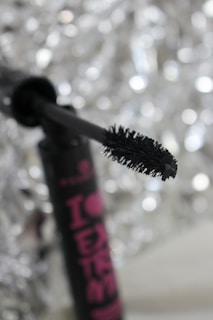 A close-up of a mascara wand with a black bristle brush in focus against a silver bokeh background. The mascara tube is partially visible with bold pink lettering.