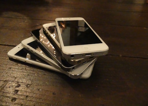 A stack of several smartphones placed on a wooden surface. The phones are assorted in colors, mainly white and black, and are arranged haphazardly. The surface has visible wood grain and is dimly lit, creating a warm ambiance.