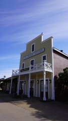 A historic building with a sign reading 'Colorado House' adorned with a wooden facade and a balcony. It has a light yellow exterior with white trim and railings. Below the main sign, there's another sign for Wells Fargo & Co. The building is set against a clear blue sky with a few clouds, and it is surrounded by some greenery.