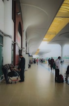A spacious, open walkway at a transportation hub with high arches and a tiled floor. People are standing and walking, some with luggage, creating a busy atmosphere. A few individuals are wearing face masks, indicating a mindful approach to health safety.