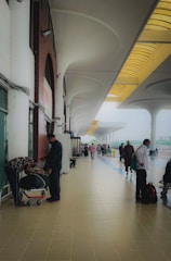 A spacious, open walkway at a transportation hub with high arches and a tiled floor. People are standing and walking, some with luggage, creating a busy atmosphere. A few individuals are wearing face masks, indicating a mindful approach to health safety.