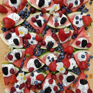 Slices of watermelon are topped with dollops of whipped cream, chia seeds, and an assortment of fresh berries including blueberries, strawberries, blackberries, and raspberries. Small white flowers are scattered among the fruit, adding a decorative touch.
