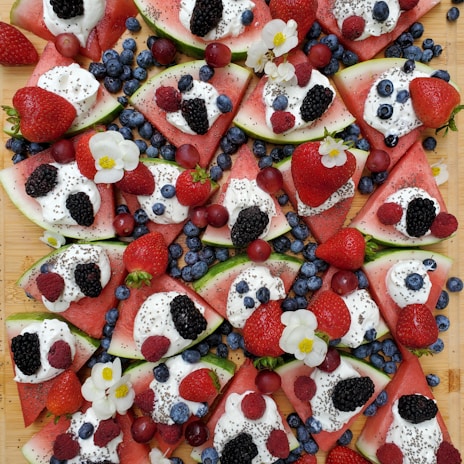 Slices of watermelon are topped with dollops of whipped cream, chia seeds, and an assortment of fresh berries including blueberries, strawberries, blackberries, and raspberries. Small white flowers are scattered among the fruit, adding a decorative touch.