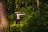 A person dressed in a traditional embroidered shirt and black pants, holding a bow and arrow in a shooting stance in a forest, surrounded by dense greenery and tall trees.