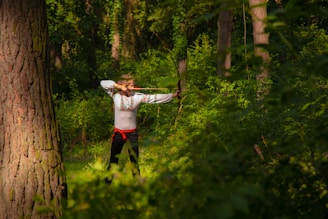 A person dressed in a traditional embroidered shirt and black pants, holding a bow and arrow in a shooting stance in a forest, surrounded by dense greenery and tall trees.