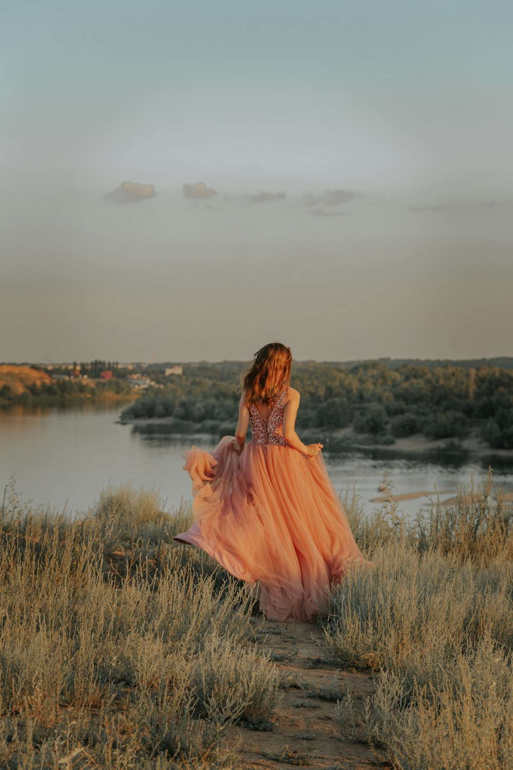 Mujer en vestido marrón de pie en el campo de hierba verde cerca del lago durante el día