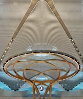 A modern architectural ceiling design featuring parallel lines and a central circular structure with golden arcs and embedded lights. A logo is suspended in the middle, creating a futuristic and symmetrical appearance.