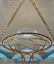 A modern architectural ceiling design featuring parallel lines and a central circular structure with golden arcs and embedded lights. A logo is suspended in the middle, creating a futuristic and symmetrical appearance.