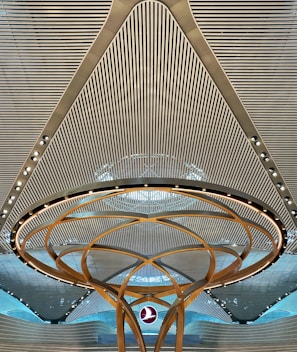 A modern architectural ceiling design featuring parallel lines and a central circular structure with golden arcs and embedded lights. A logo is suspended in the middle, creating a futuristic and symmetrical appearance.