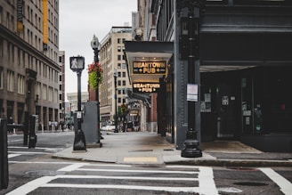 A quiet urban street featuring a pub sign with 'Beantown Pub' illuminated. Surrounding it are tall, multi-story buildings with classic architectural designs. The scene includes streetlights, crosswalks, and scattered pedestrians in the background. Flower baskets add a touch of nature to the otherwise industrial setting.