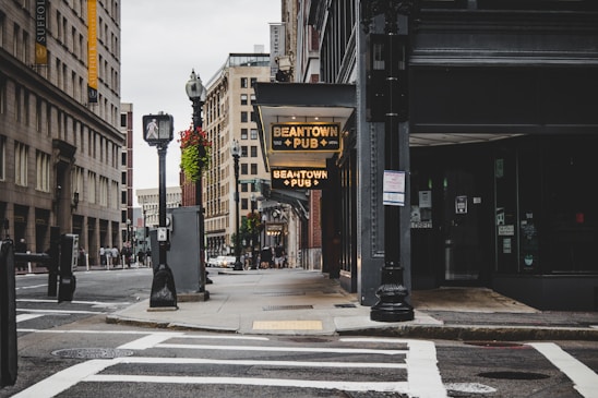 A quiet urban street featuring a pub sign with 'Beantown Pub' illuminated. Surrounding it are tall, multi-story buildings with classic architectural designs. The scene includes streetlights, crosswalks, and scattered pedestrians in the background. Flower baskets add a touch of nature to the otherwise industrial setting.