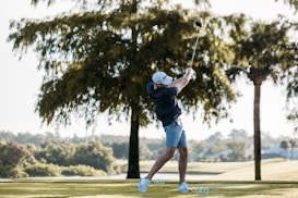 A person is captured mid-swing on a golf course, surrounded by lush greenery and large trees. The golfer is wearing a cap, navy blue shirt, and light blue shorts.