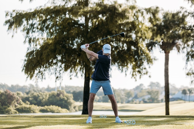 A person in sports attire is preparing to swing a golf club on a lush green golf course. The individual wears a dark shirt, light shorts, and a cap. Numerous golf balls are scattered near their feet, and a large tree with dense foliage stands behind. Sunlight filters through the trees, creating dappled shadows on the ground.