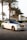 A luxury white Rolls Royce car is parked on a cobblestone road lined with tall palm trees. The car has a sleek design with a glossy black roof and shiny chrome accents. The background includes neatly trimmed bushes and a clear sky, suggesting a warm climate.