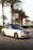 A luxury white Rolls Royce car is parked on a cobblestone road lined with tall palm trees. The car has a sleek design with a glossy black roof and shiny chrome accents. The background includes neatly trimmed bushes and a clear sky, suggesting a warm climate.
