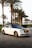 A luxury white Rolls Royce car is parked on a cobblestone road lined with tall palm trees. The car has a sleek design with a glossy black roof and shiny chrome accents. The background includes neatly trimmed bushes and a clear sky, suggesting a warm climate.
