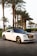 A luxury white Rolls Royce car is parked on a cobblestone road lined with tall palm trees. The car has a sleek design with a glossy black roof and shiny chrome accents. The background includes neatly trimmed bushes and a clear sky, suggesting a warm climate.