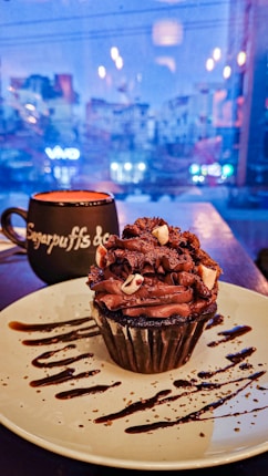 A chocolate cupcake with rich frosting topped with chocolate shavings and small pieces of white chocolate sits on a plate decorated with chocolate drizzle. In the background, a cup with a saucer is visible, featuring a name or brand on it. The scene is set indoors with a window view of a cityscape that is slightly blurred and illuminated by streetlights.