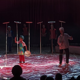 A circus performer dressed as a chef and a young participant are engaged in a plate-spinning act. Several plates are spinning atop long poles, while four individuals in uniform stand in the background observing. The ground is covered with a shiny red material, and the scene is dimly lit, creating a dramatic atmosphere.